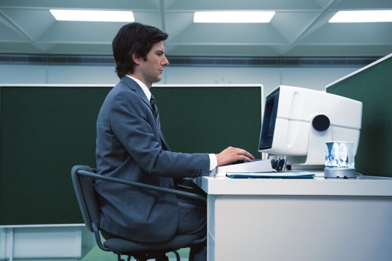 Man in profile, white, short and straight hair, sitting in front of an old-looking computer, between green office partitions, with artificial lights on the ceiling.