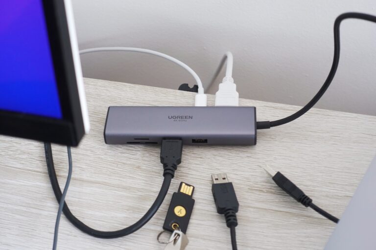 Detalhe de um hub USB-C sobre um mesa, atrás de um monitor parcialmente visível, com vários cabos conectados e algumas pontas USB-A ao lado, soltas.