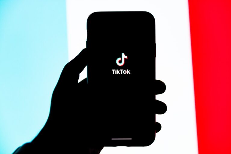 Silhueta de uma mão segurando um celular com o logo do TikTok na tela. Ao fundo, listas azul, branca e vermelha.