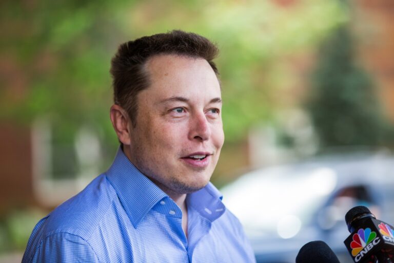 Elon Musk, vestindo camisa azul, com microfones à sua frente.