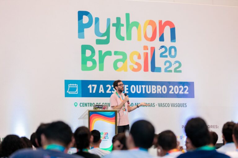 Homem branco, em um palco, com cabeças desfocadas em primeiro plano. Ao fundo, um painel enorme com os dizeres “Python Brasil 2022” em letras coloridas.