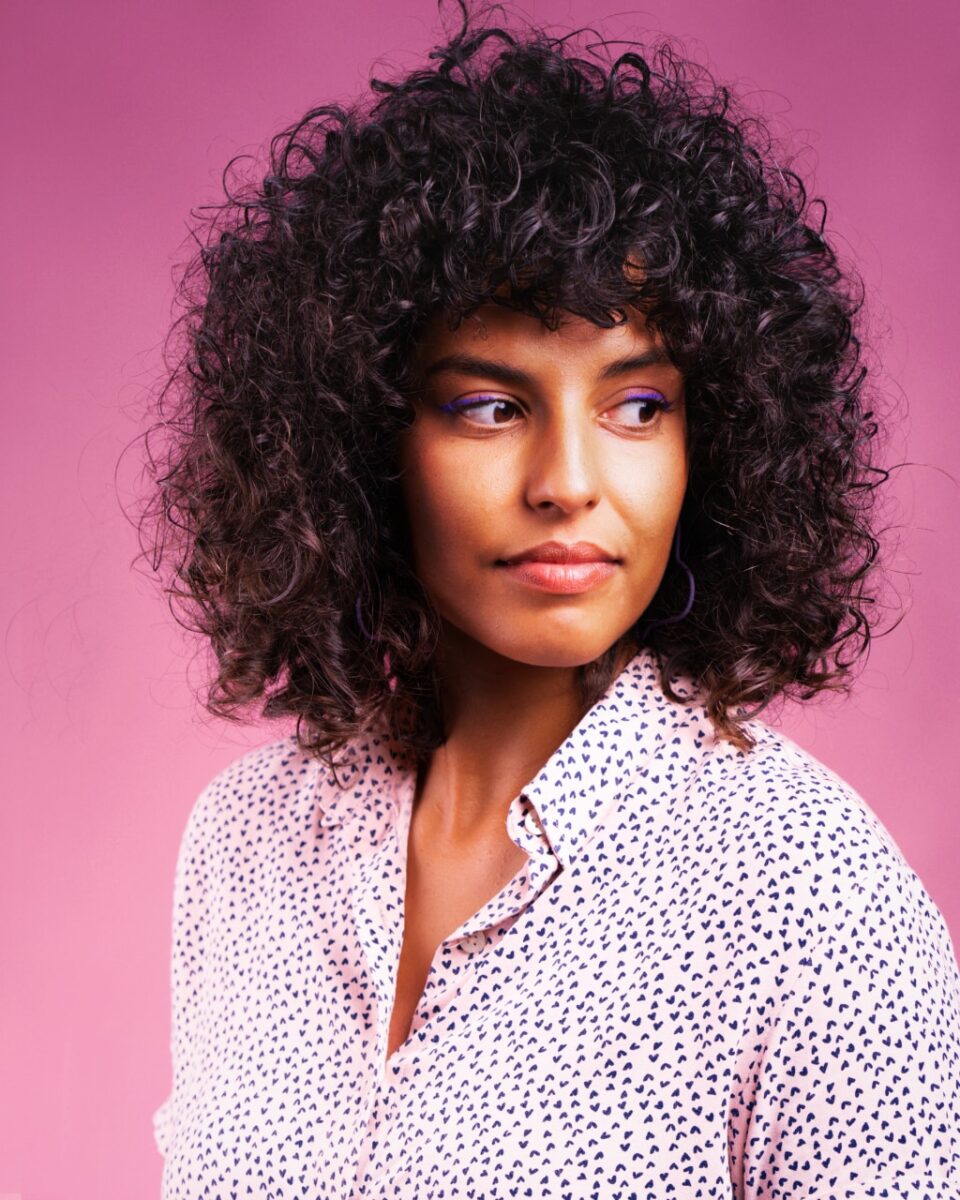 Retrato de Nara Oliveira: mulher preta, de cabelos cacheados, vestindo uma camisa clara com bolinhas escuras, olhando à sua esquerda.