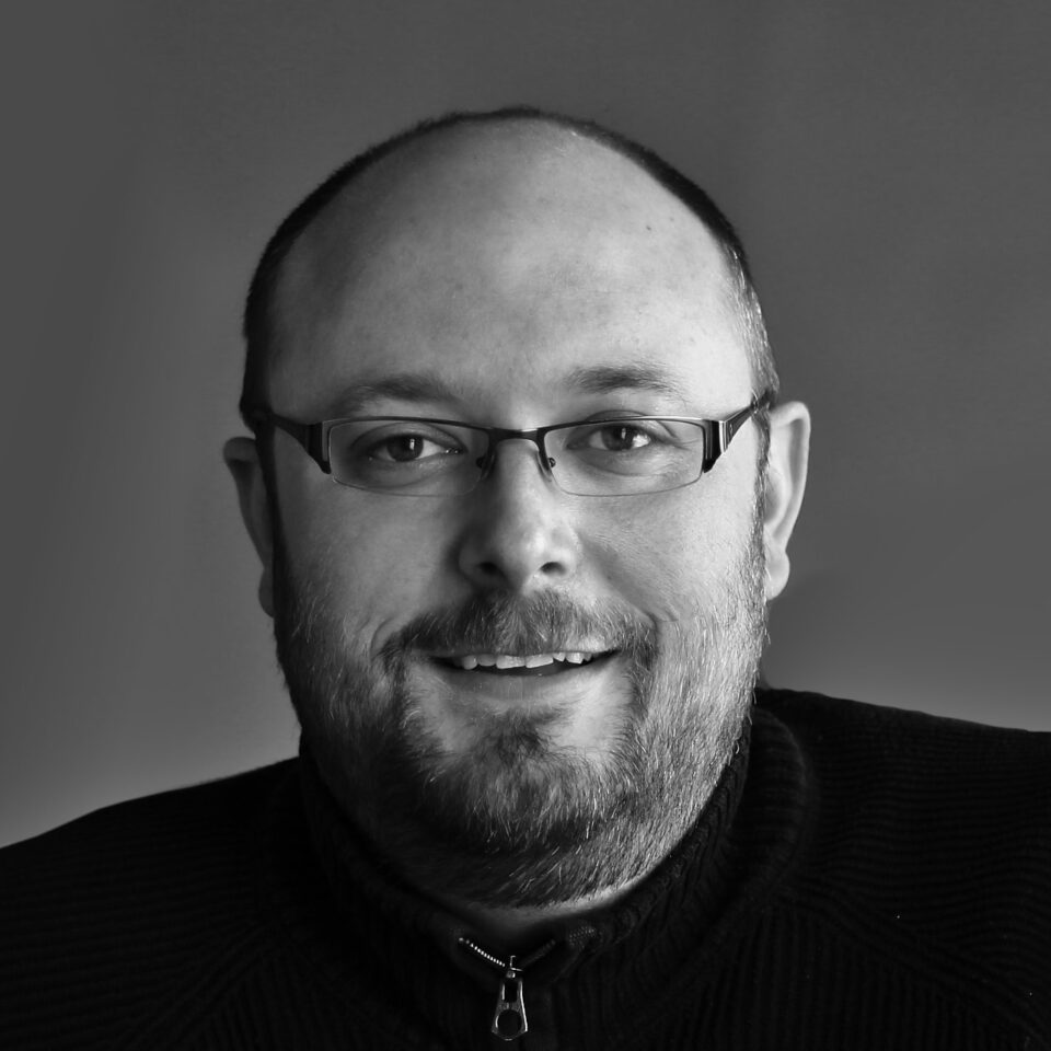 Retrato em preto e branco de Ryan Gorley: homem branco, calvo, com barba curta e óculos de grau, vestindo uma camiseta ou camisa escura.