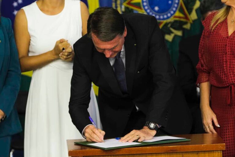 Jair Bolsonaro, de pé, inclinado sobre uma mesa, assinando um papel. Ao seu redor, três mulheres (sem mostrar os rostos delas).
