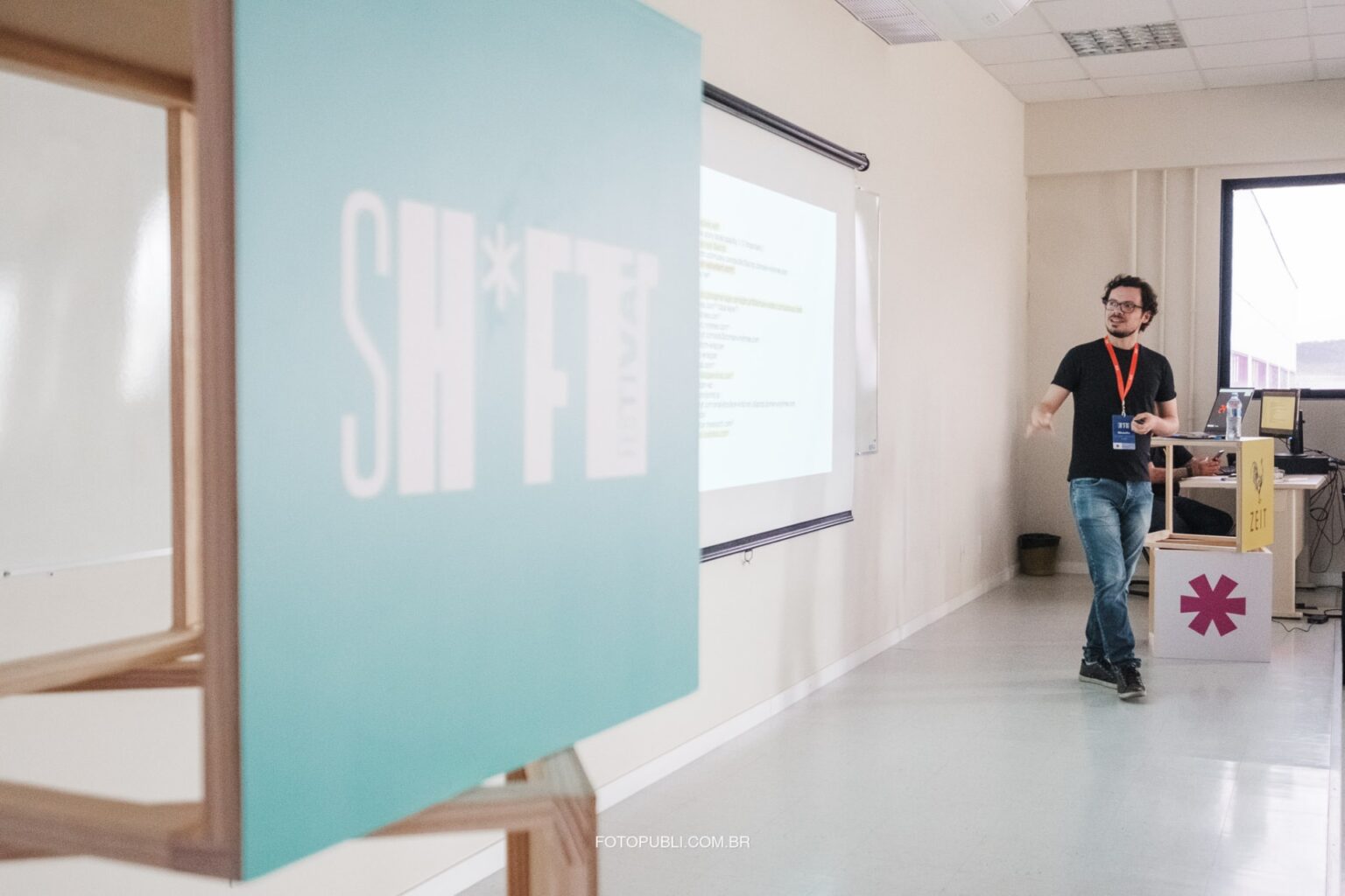 Em primeiro plano, desfocada, uma placa do SH*FT; ao fundo, em foco, Rodrigo Ghedin (homem branco, de camiseta preta e calça jeans), de pé, falando enquanto olha para uma lousa branca.