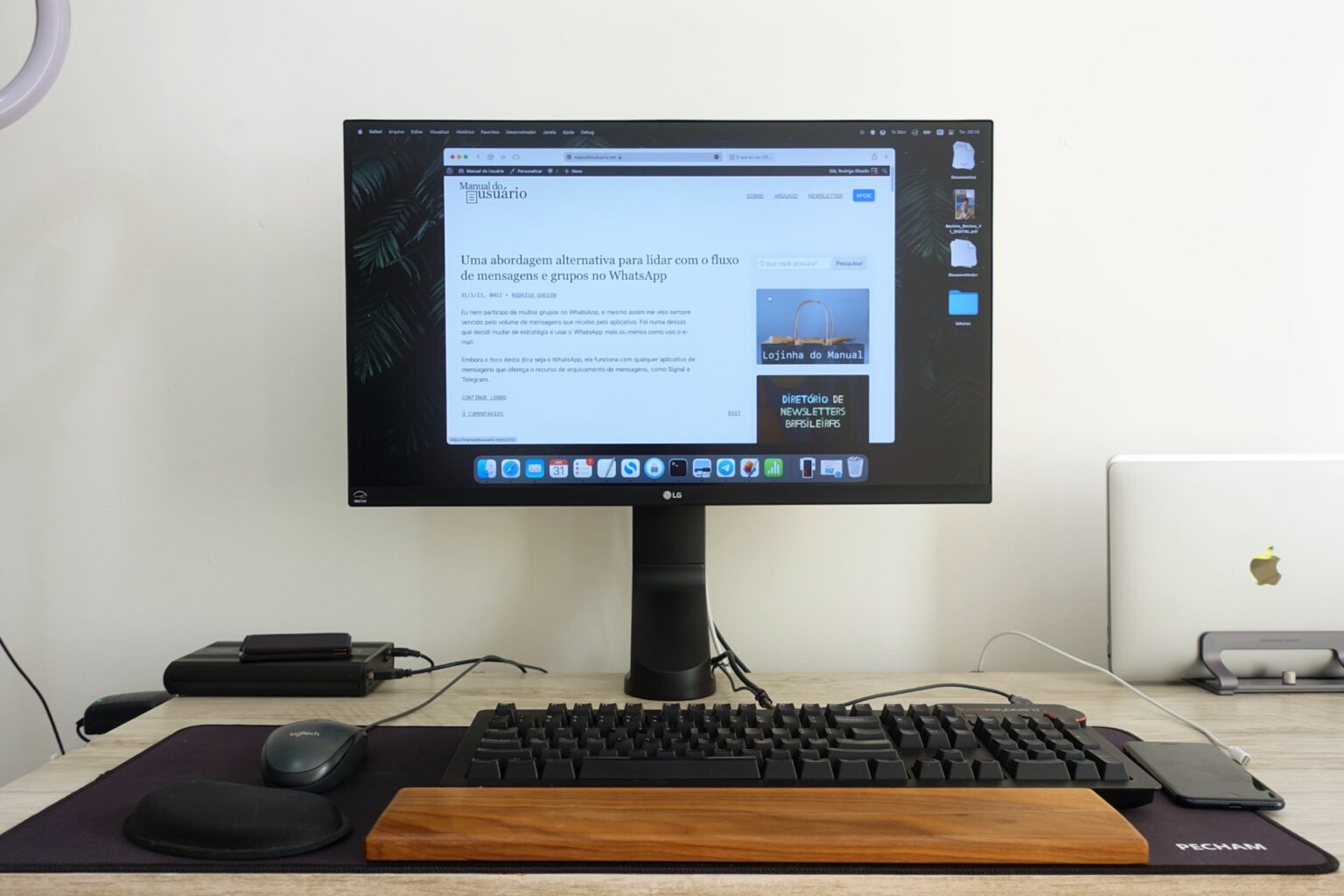 Foto frontal de uma mesa de trabalho, com o monitor ligado mostrando o Safari aberto no Manual do Usuário. Embaixo, um mousepad grande com mouse, teclado e apoios para os pulsos, um de madeira, outro de espuma/preto. Ao fundo, uma carteira sobre um case de HD externo. À direita, MacBook Air fechado, de pé.