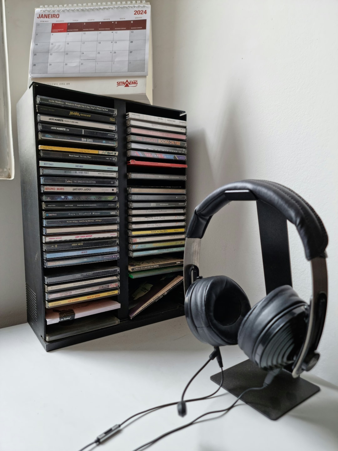 Headset em um suporte em primeiro plano e, ao fundo, dois porta-CDs verticais quase cheios de CDs.