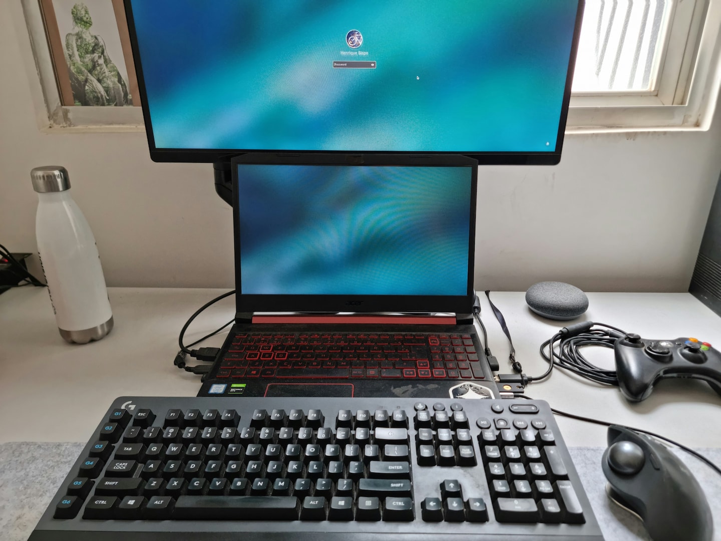 Detalhe da mesa, mostrando teclado, mouse, notebook aberto e uma tela grande.