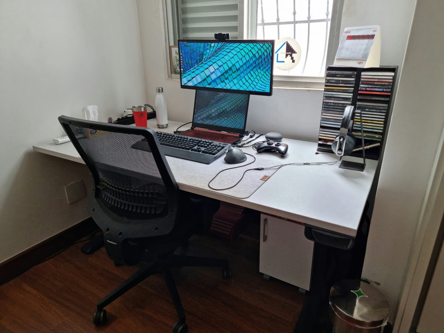 Vista geral da mesa de trabalho do Henrique Bispo, contra uma janela aberta, num canto do cômodo, com uma cadeira.