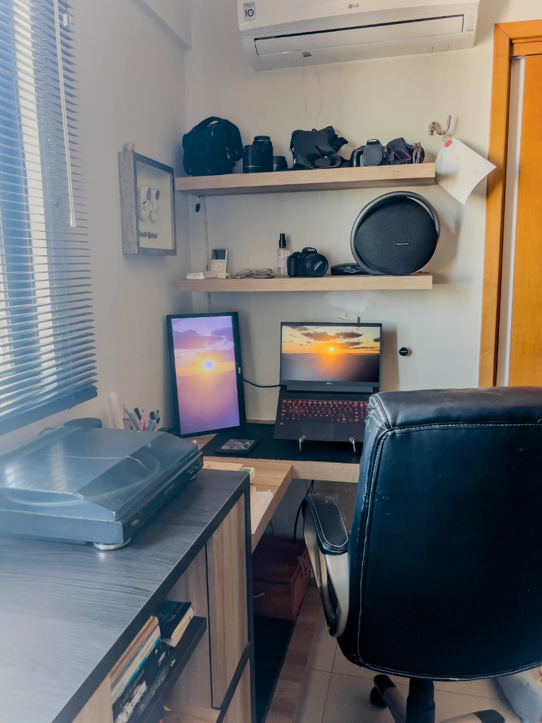 Mesa de canto, com notebook e monitor externo, uma vitrola ao fundo e caixa de som, câmeras e iPods nas prateleiras superiores.
