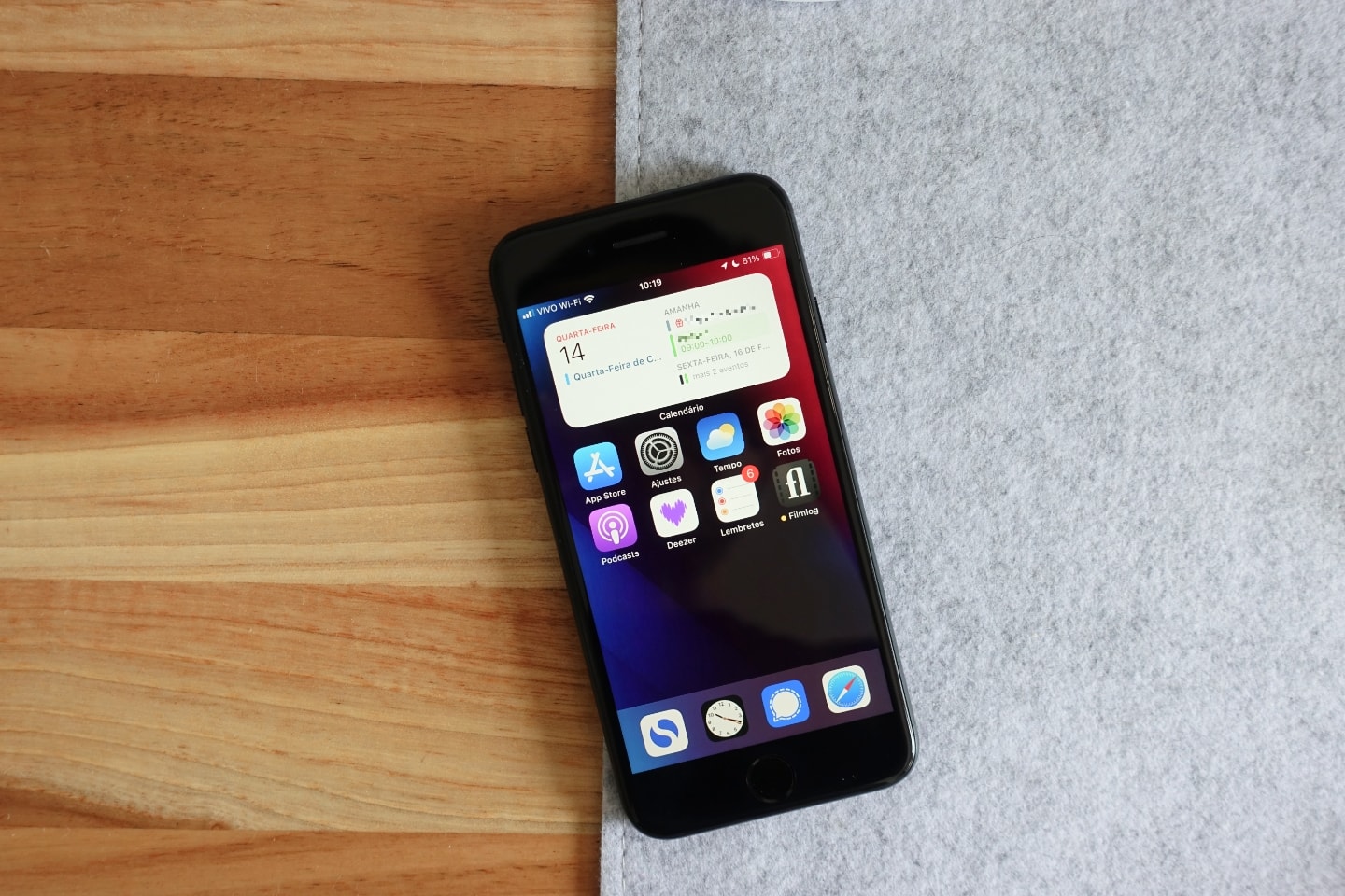 Foto de cima de um iPhone SE, na divisa entre a mesa de madeira e deskmat claro de feltro.