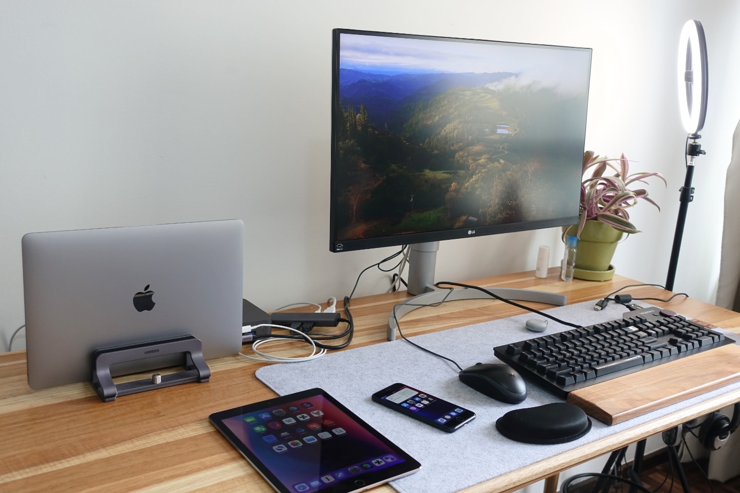 Foto na diagonal de uma mesa de trabalho, com monitor grande, celular, tablet e acessórios espalhados por cima.