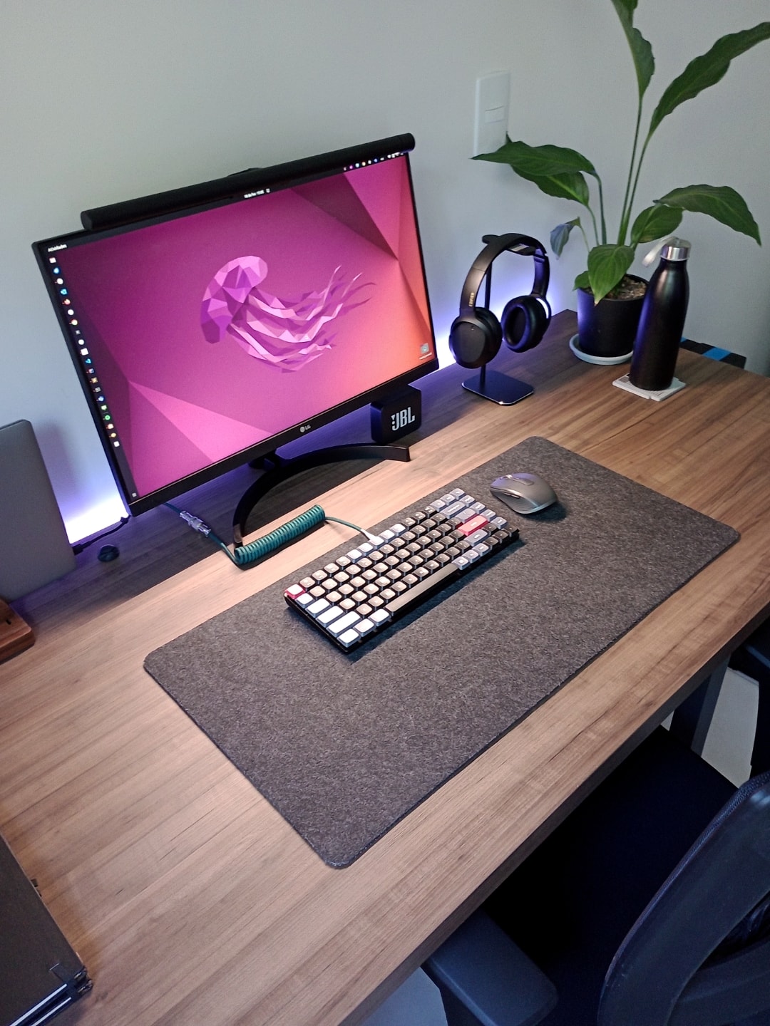 Foto na diagonal da mesa do Leonardo, mostrando monitor, teclado, mouse, fones de ouvido, garrafa d'água e planta.