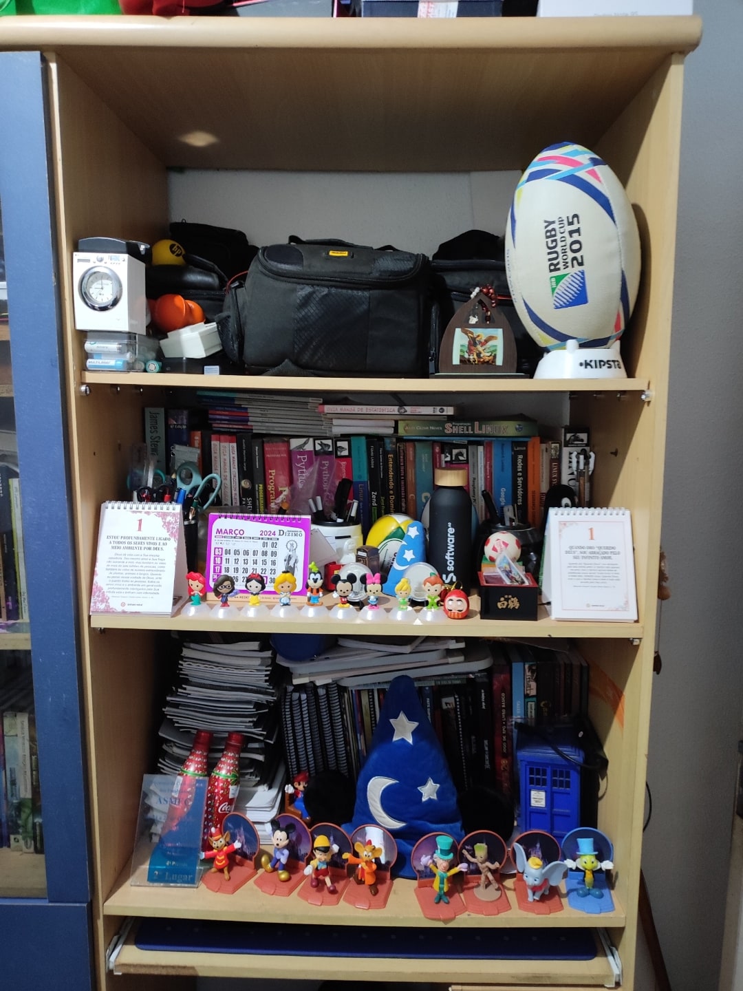 Foto frontal de uma estante, com bola de futebol americano, livros e bonecos.