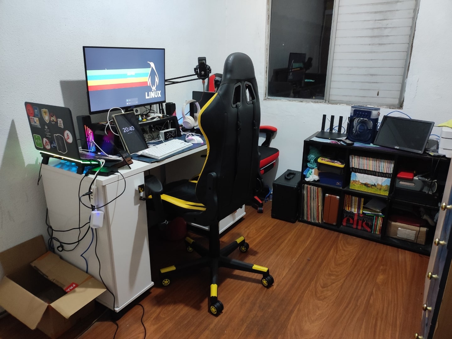 Foto lateral da mesa de trabalho do Fábio, com dois notebooks e monitor com papel de parede Linux, e uma pequena mesa e janela ao fundo.