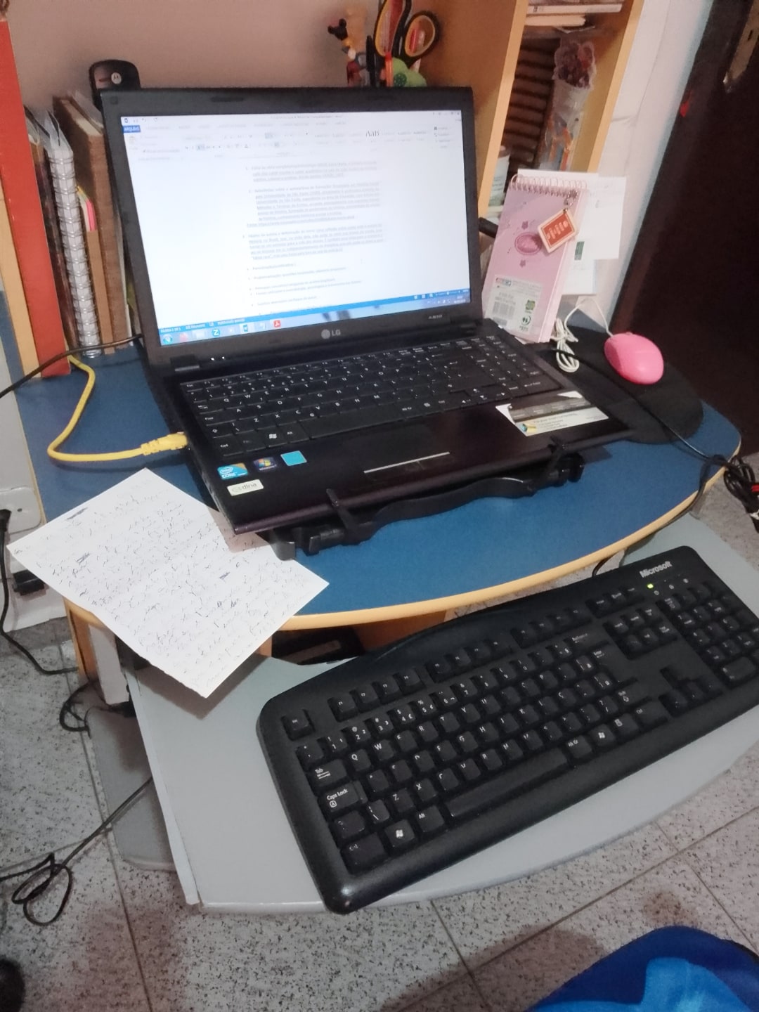 Foto aproximada da mesa da Keli, mostrando o notebook e o teclado à parte, numa gaveta embaixo da mesa.