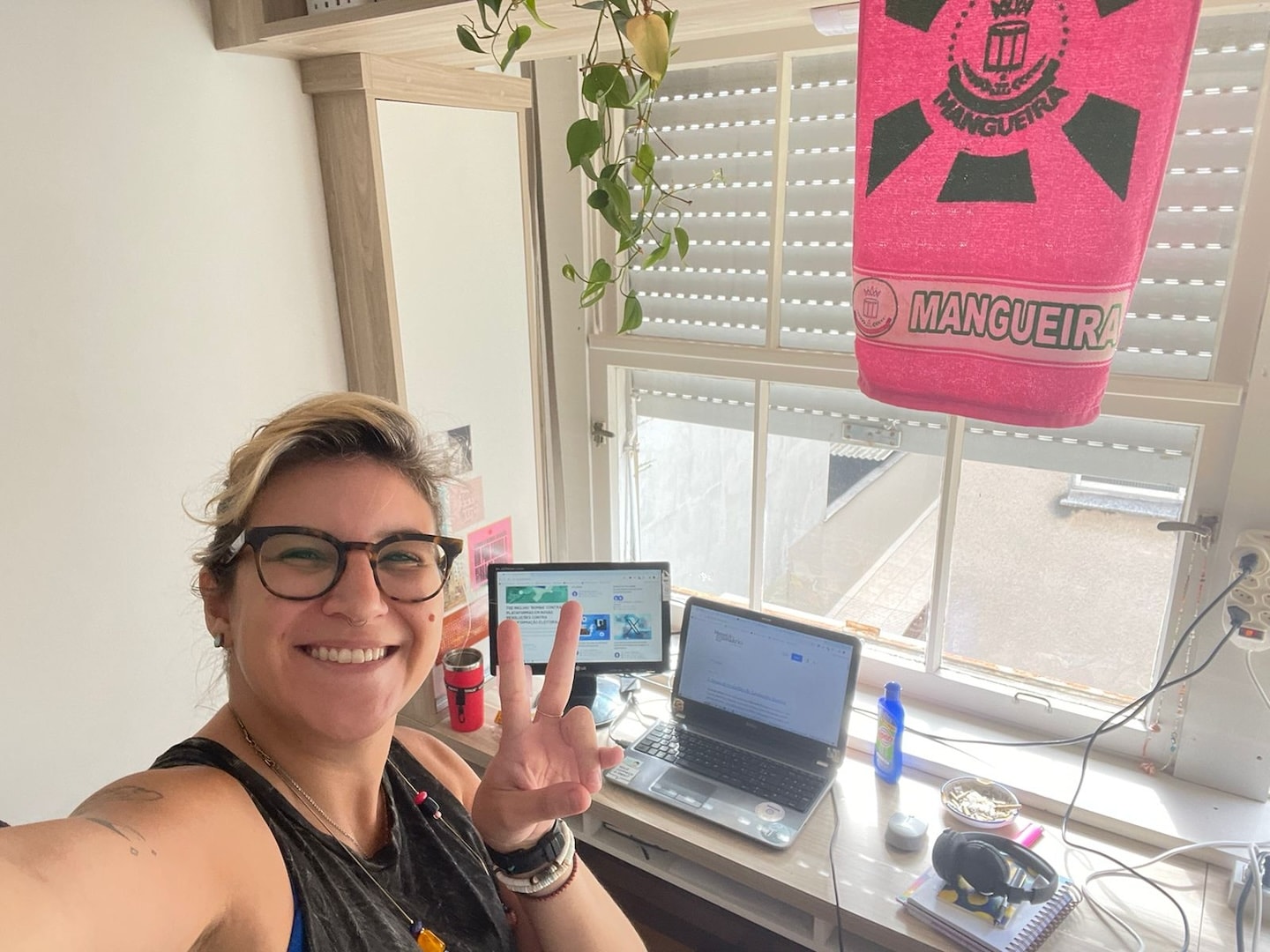 Selfie de mulher loira, de óculos, fazendo um “V” com a mão. Ao fundo, notebook e monitor em uma bancada contra uma janela semi-aberta.