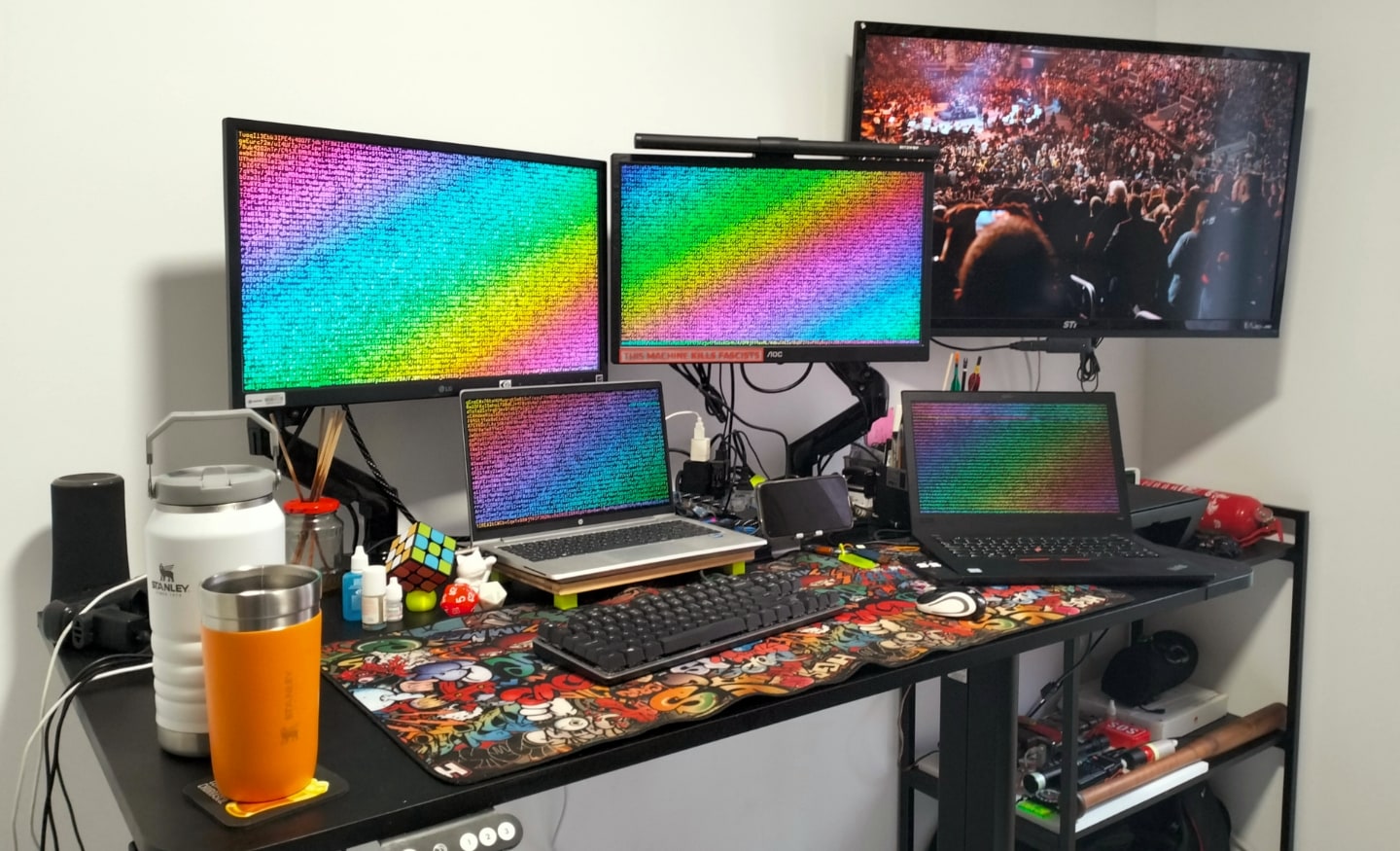 Foto lateral da mesa do Carlos, com dois monitores, dois notebooks, TV e acessórios.