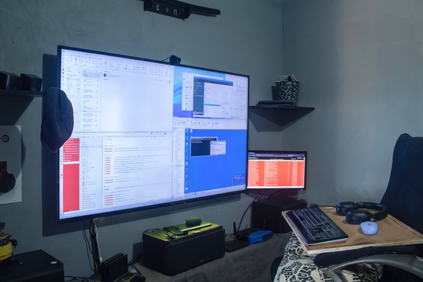 Foto de lado, de uma TV grande e um monitor menor, com uma cadeira com mouse e teclado sobre uma placa de madeira em cima de uma almofada.