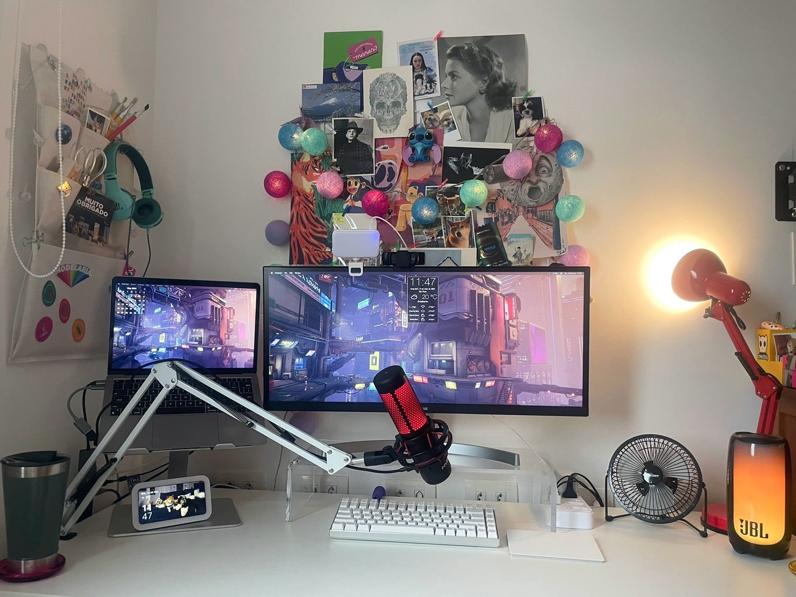 Foto frontal da mesa da Ieda, com monitor, acessórios e um painel de colagem na parede atrás.