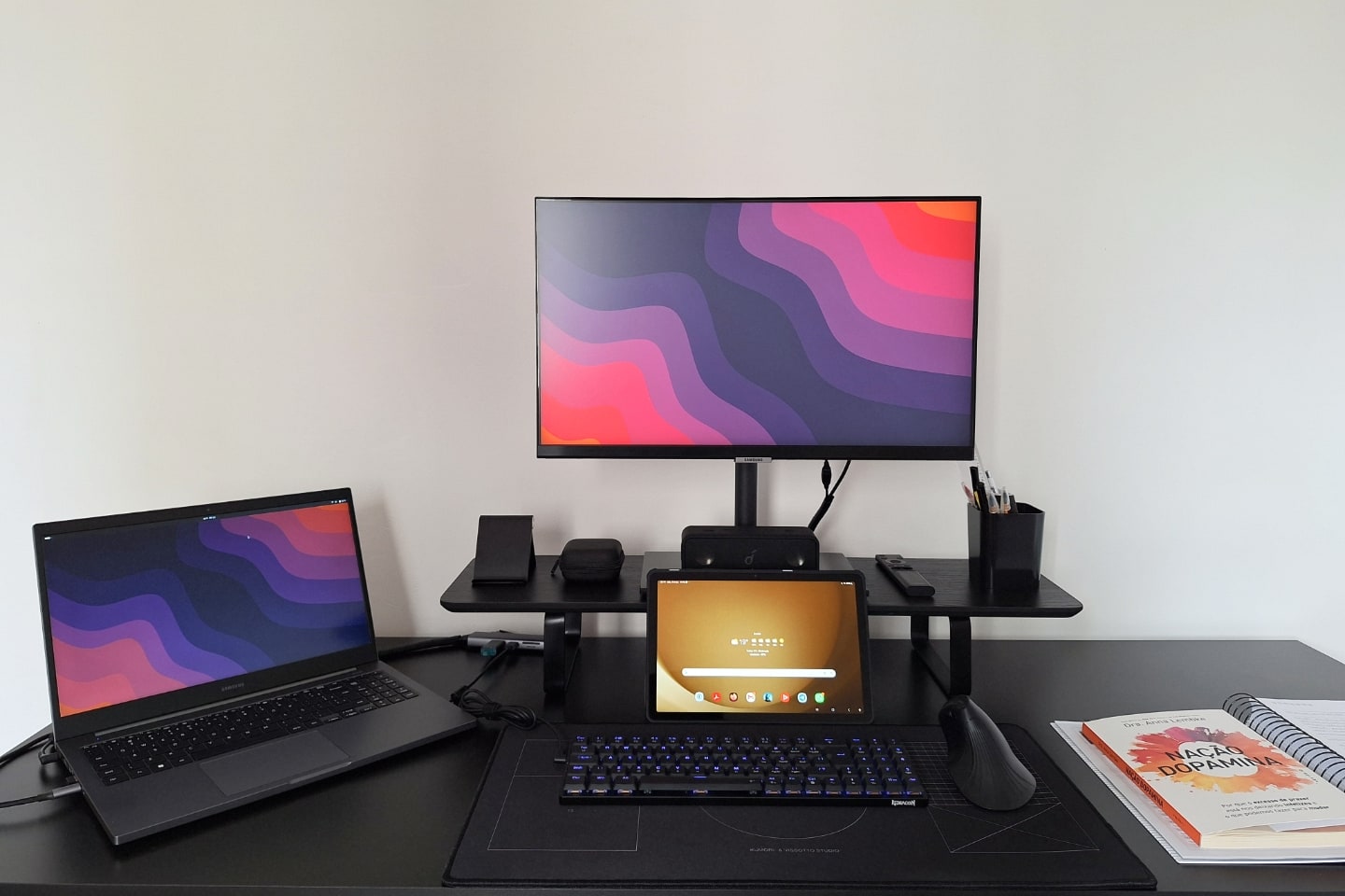 Foto frontal da mesa do Eduardo, com um notebook à esquerda, monitor e tablet centralizados, alguns acessórios e um livro.