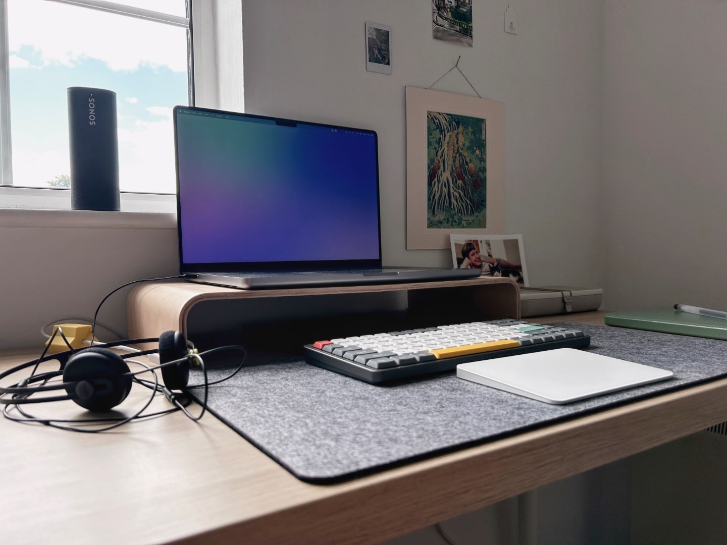 Foto na diagonal de uma mesa em frente a uma janela com notebook, teclado e trackpad, e fones de ouvido à esquerda.