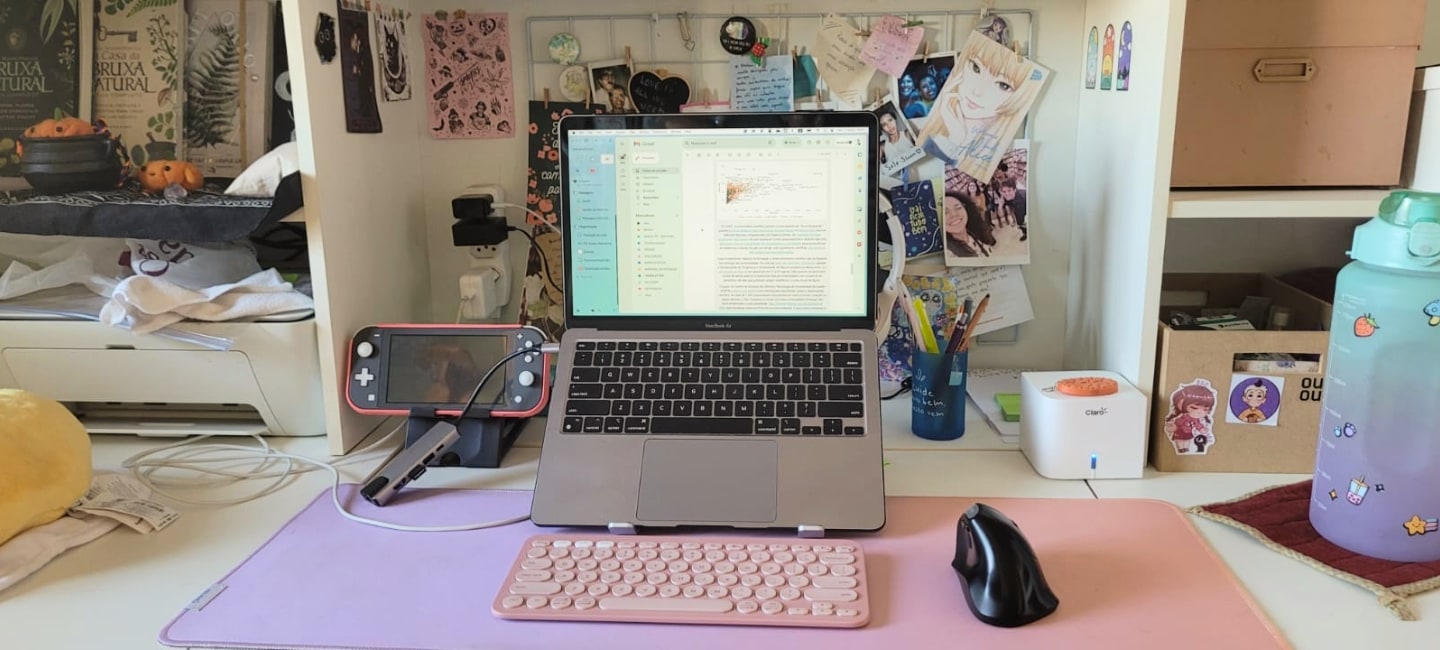 Foto frontal de uma mesa de trabalho, com um notebook aberto elevado, teclado e mousepad rosas, mouse vertical e Nintendo Switch Lite, colagens na parede e outros acessórios.