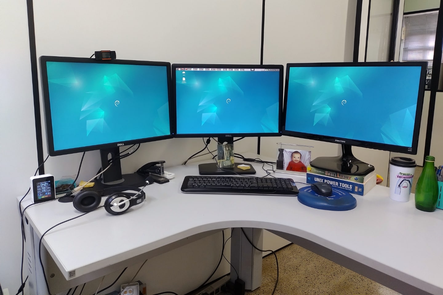 Mesa em “L” com três monitores ligados, exibindo um papel de parede azul do Debian.