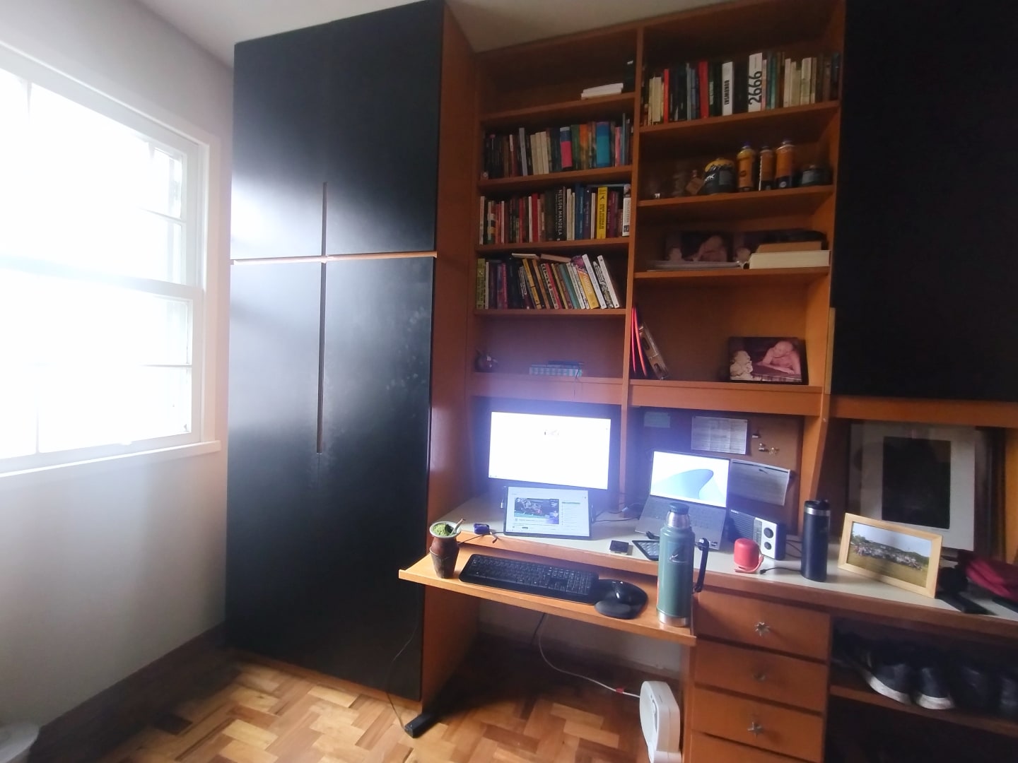 Foto aberta de um móvel de madeira grande, que cobre toda a parede, com mesa de computador embutida, estante de livros e armário. À esquerda, janela com iluminação vinda de fora.