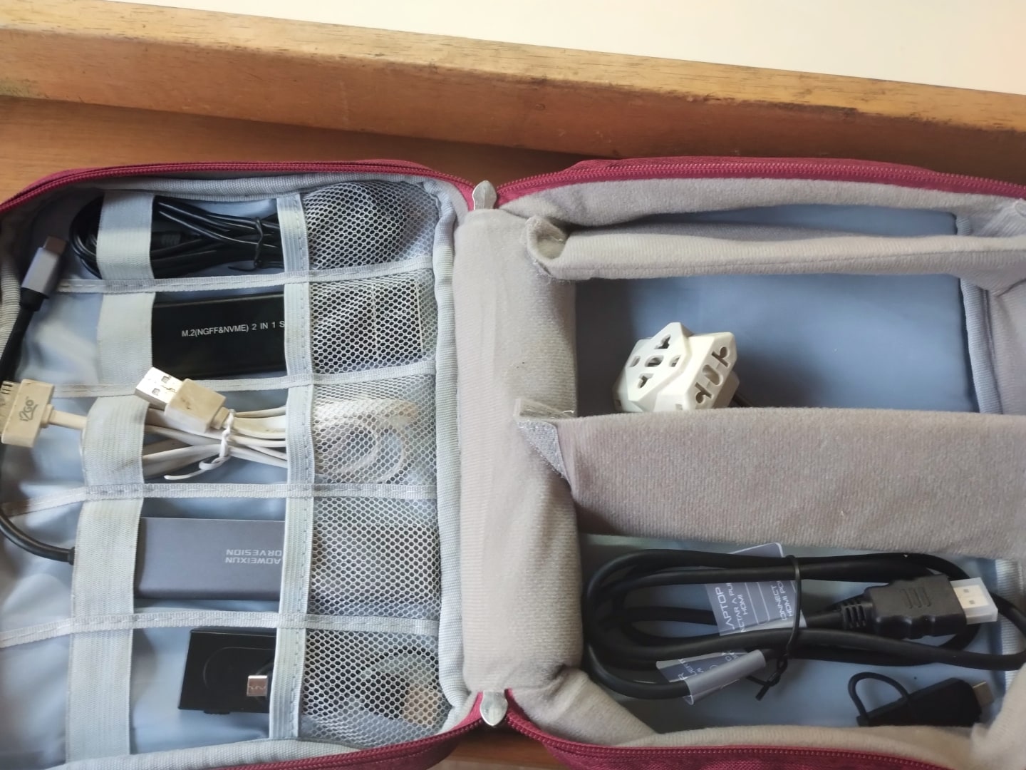 Organizador de cabos aberto, com tecido interno e divisórias cinzas, alguns cabos e adaptadores.