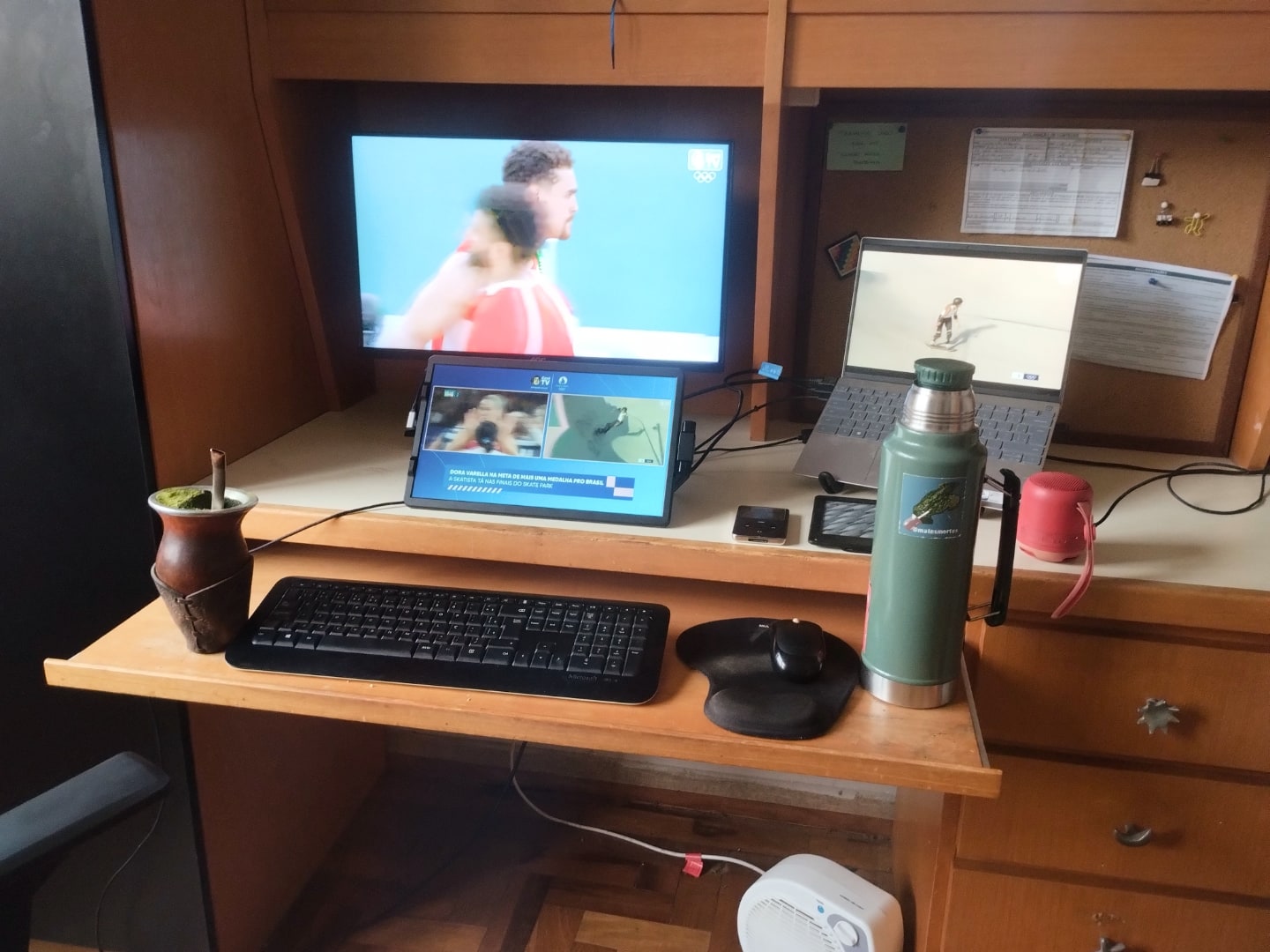 Mesa de trabalho com notebook à direita, dois monitores alinhados na vertical, teclado, mouse, cuia de chimarrão e garrafa térmica.