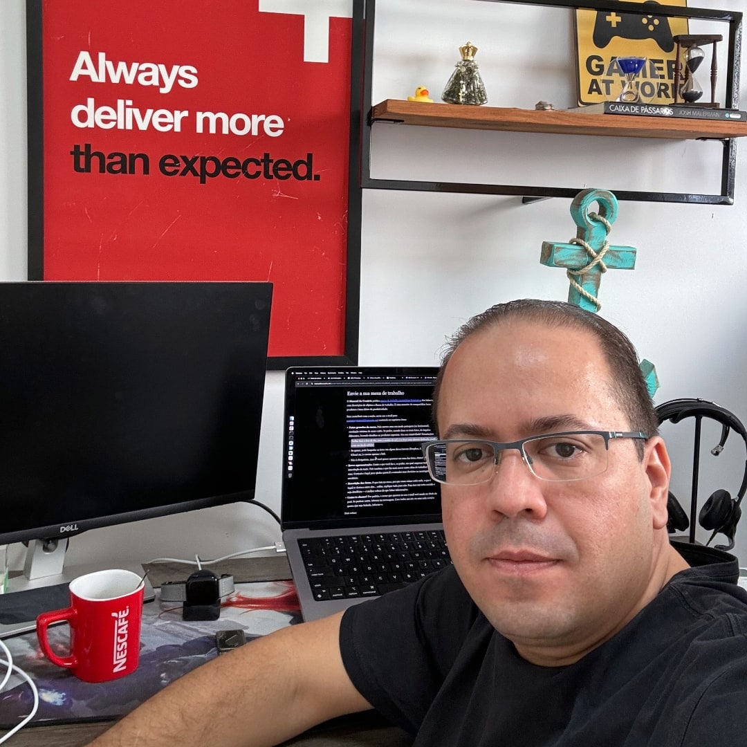 Selfie de homem branco, de óculos de grau e cabelo curto, vestindo camiseta preta, à frente de computador, monitor, quadro e outros elementos decorativos.