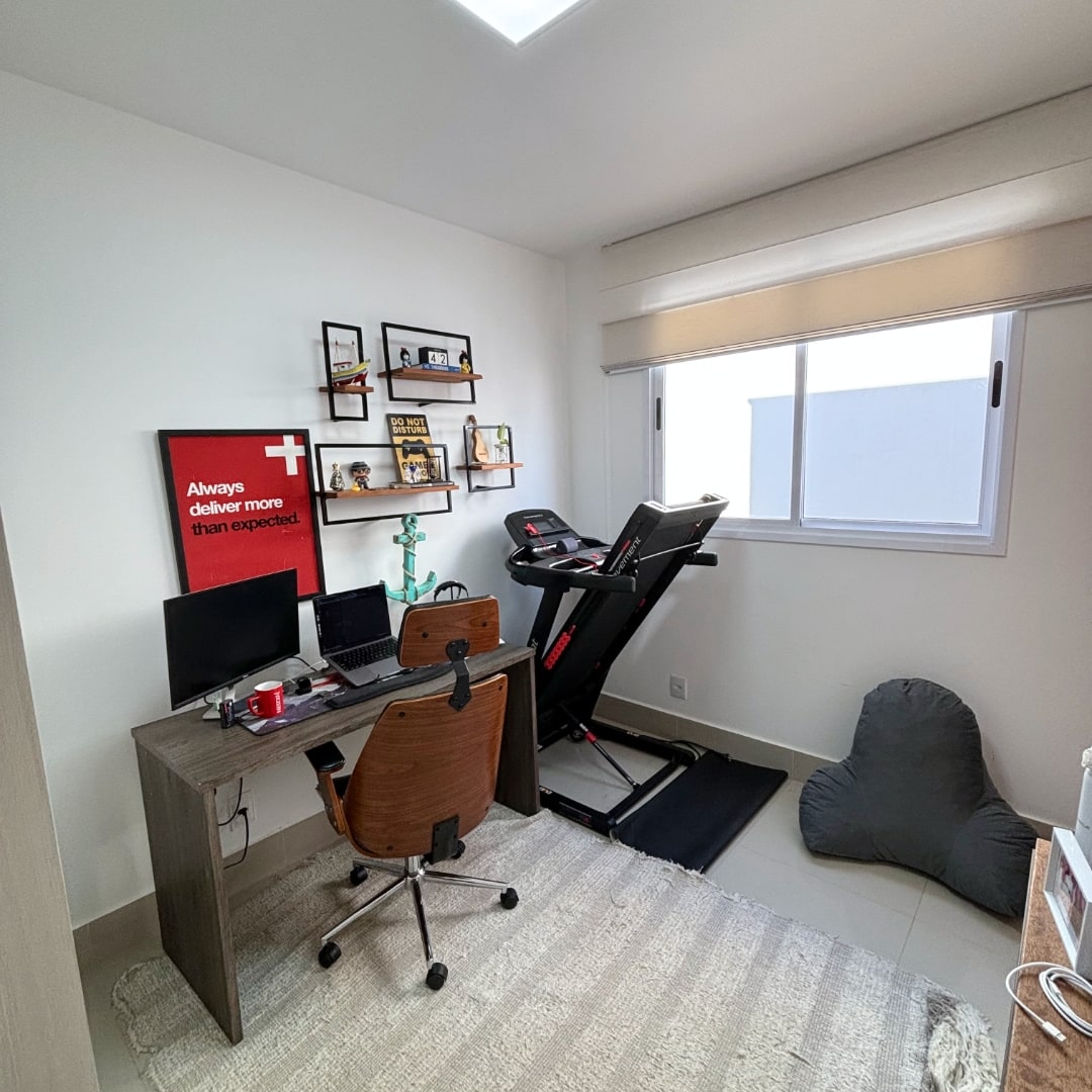 Visão geral da mesa de trabalho do Vitor, com prateleiras decorativas, uma esteira e uma janela do lado direito.