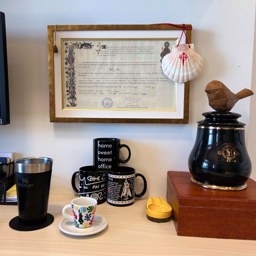 Detalhe da mesa, com canecas, copos e certificado do Caminho de Santiago da Compostela na parede.