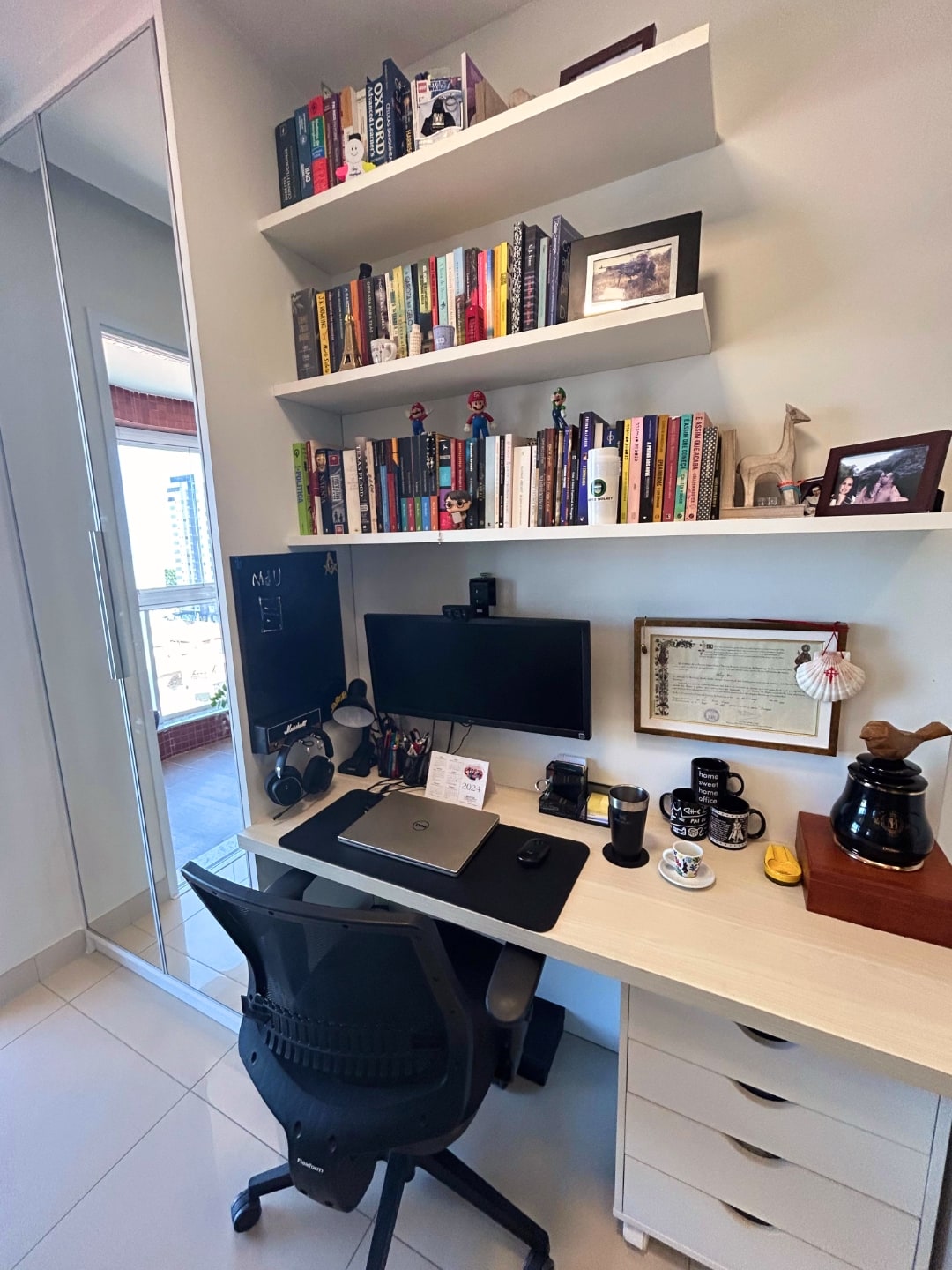 Foto em retrato da mesa do Wolney, mostrando a cadeira e mais prateleiras, e um espelho à esquerda.