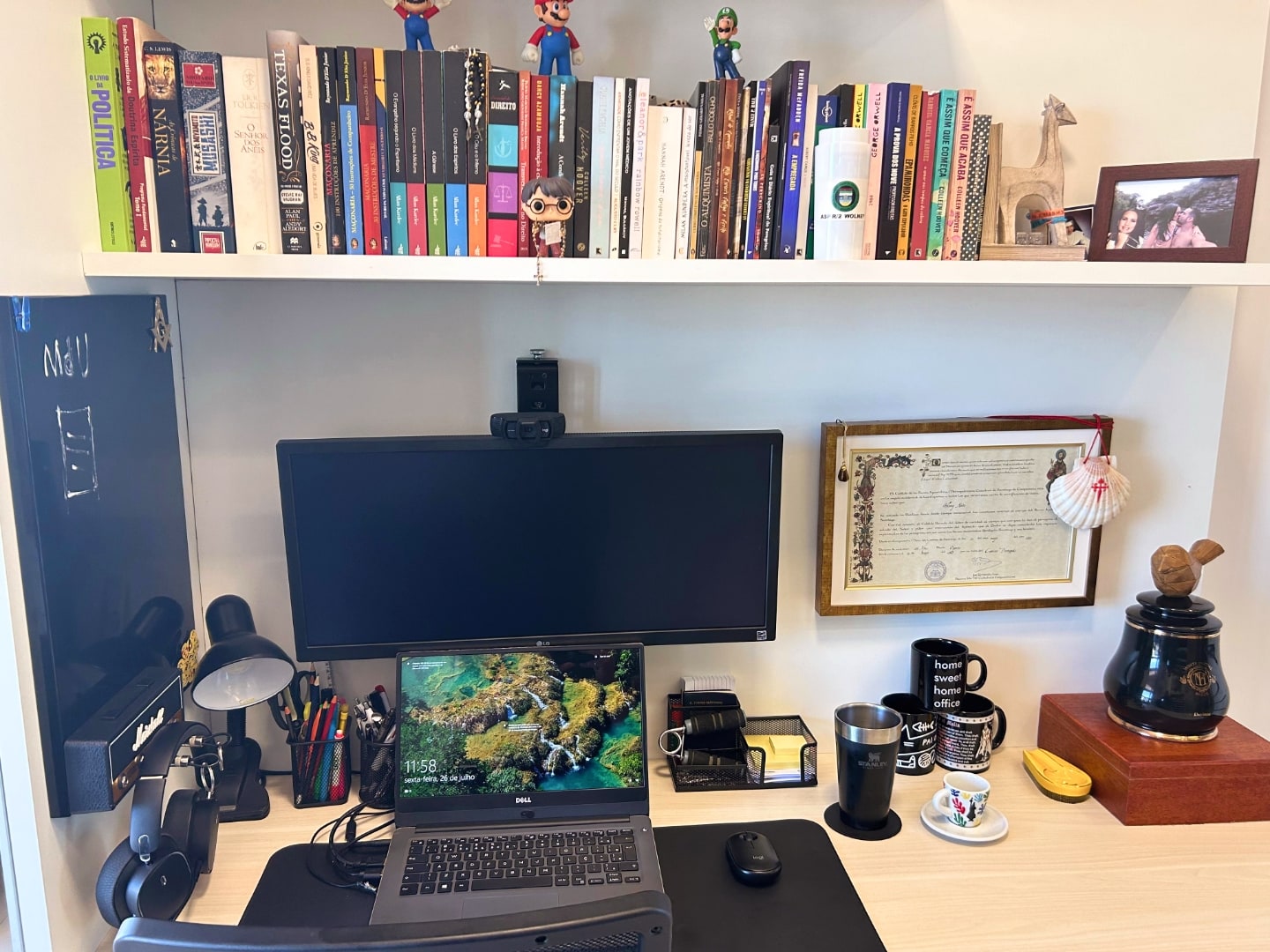 Foto frontal da mesa do Wolney, com computador, prateleira de livros e diversos objetos sobre a mesa.