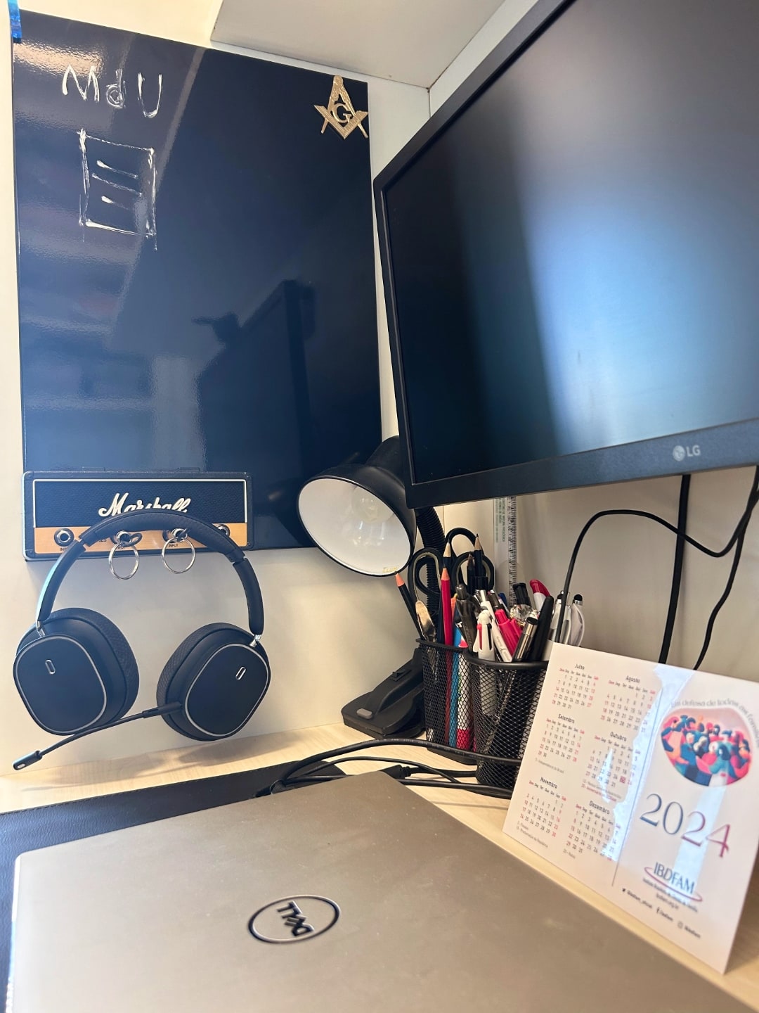 Detalhe da mesa, mostrando lousa, fones de ouvido, luminária e monitor.