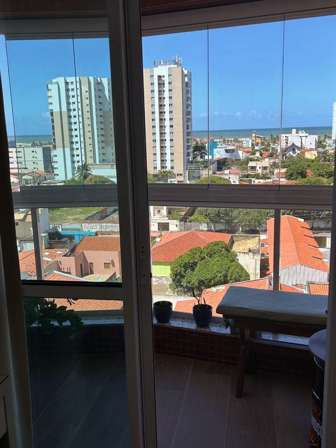 Vista da varanda, com dois prédios, casas, árvores e o mar ao fundo.