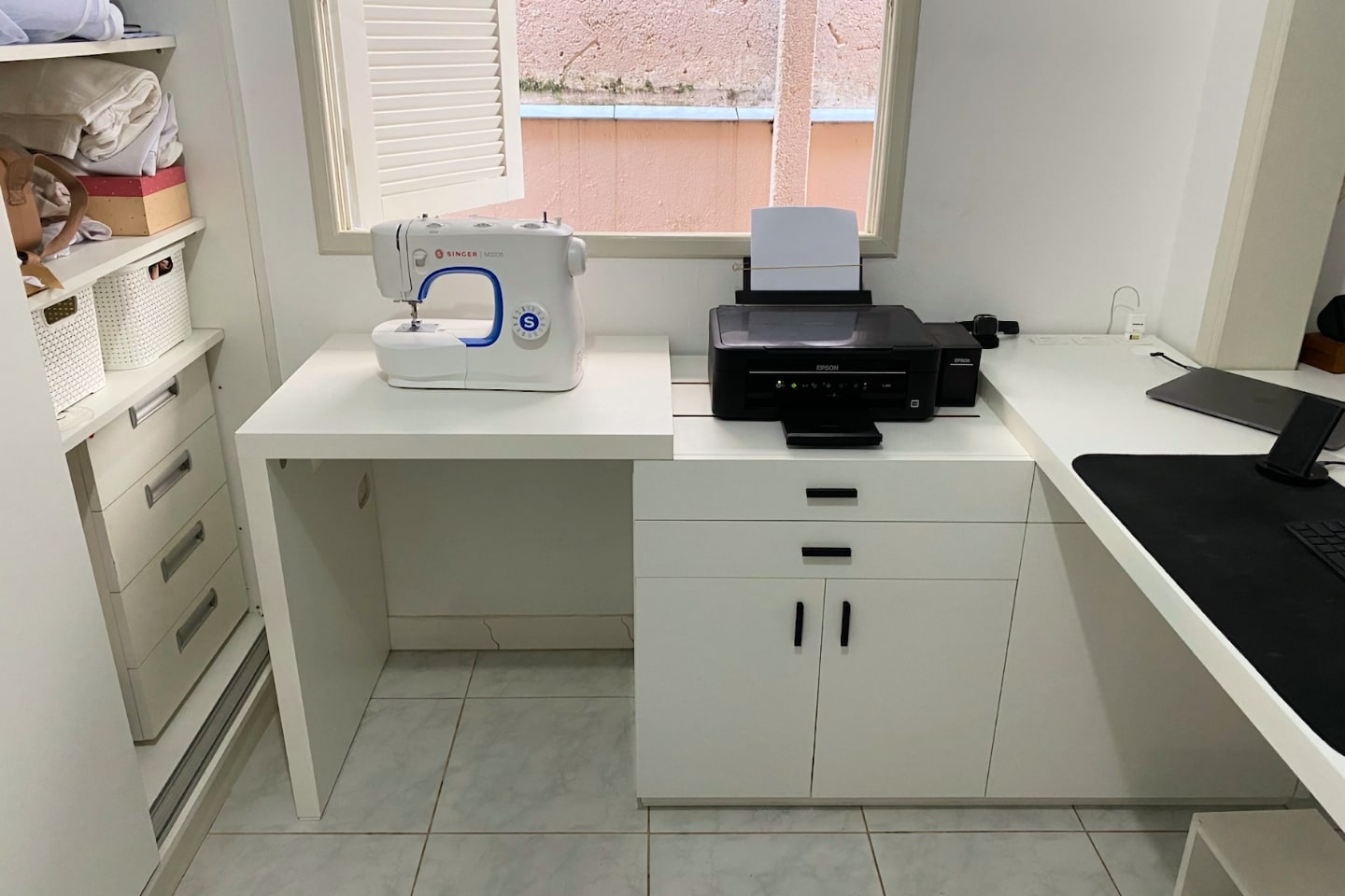 Impressora e máquina de costura sobre mesa/bancada em frente a uma janela aberta.