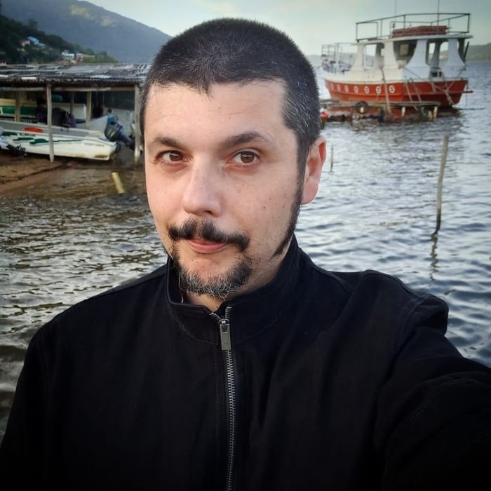 Homem branco, de cabelo curto e barba, em frente a um rio com alguns barcos.