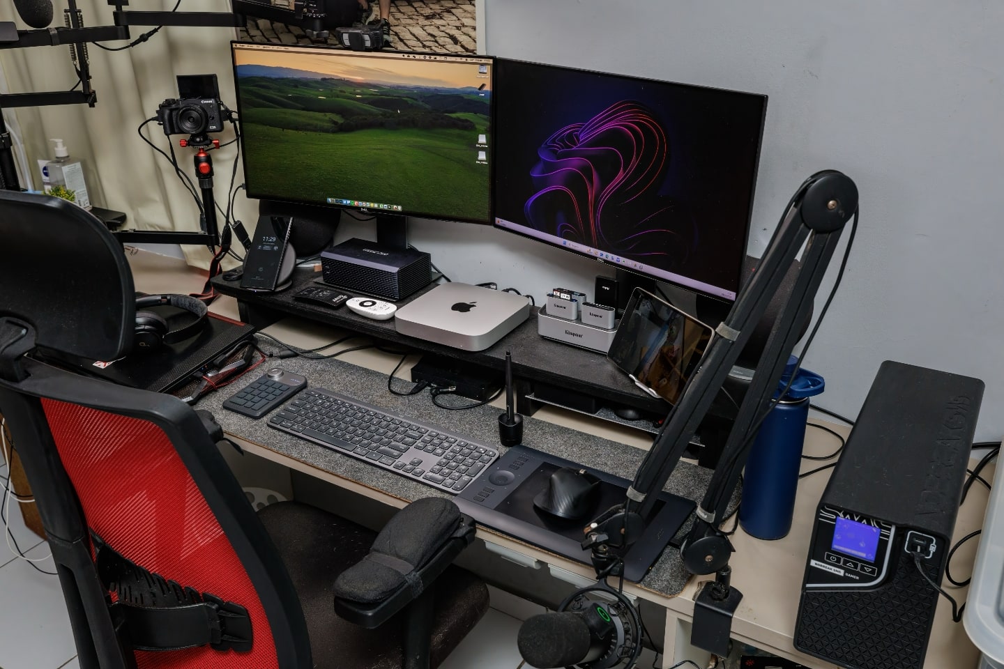 Foto na diagonal da mesa do Albert, com dois monitores, teclado e mouse, computadores e outros acessórios.