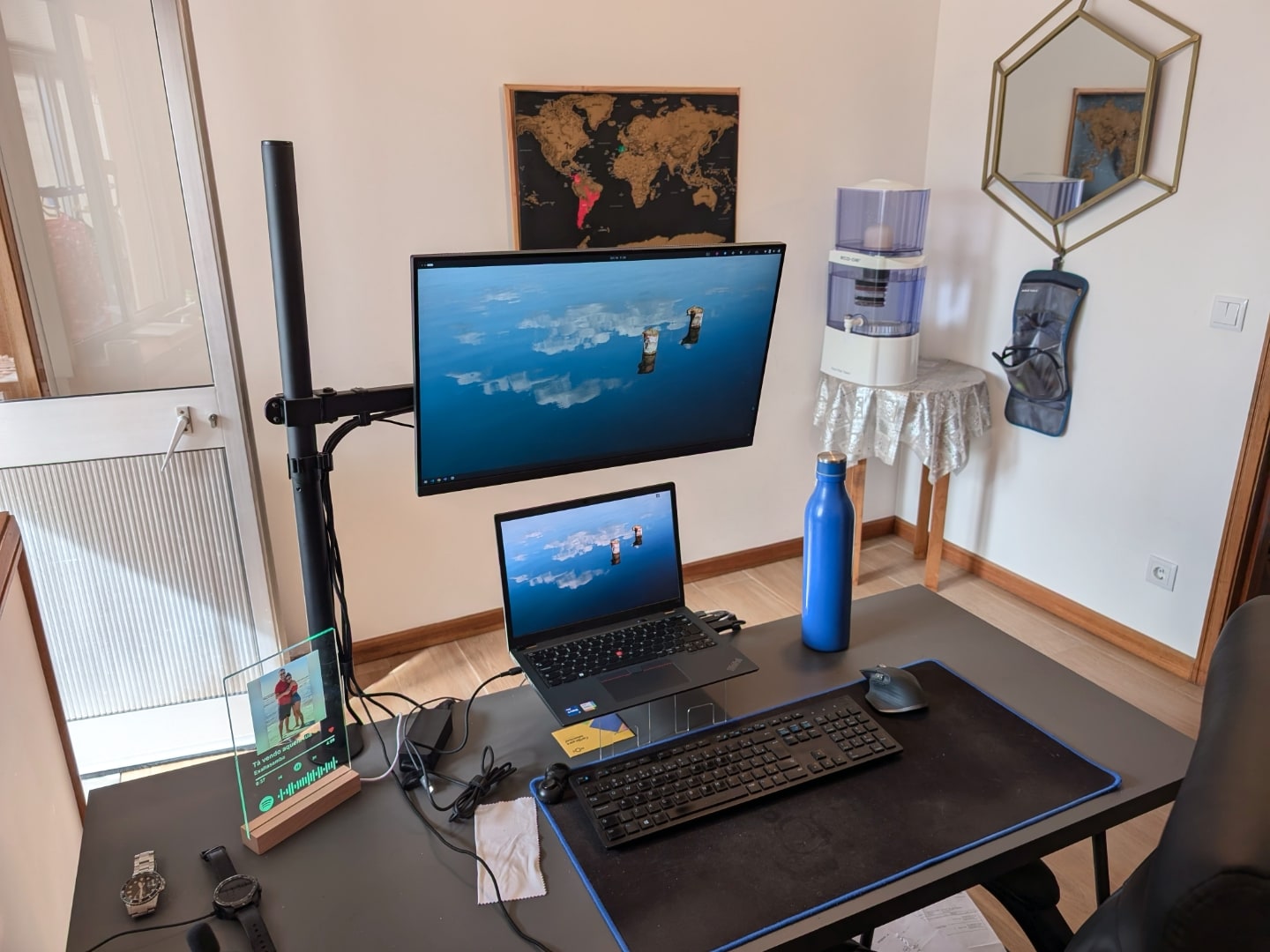 Mesa de trabalho com notebook, monitor suspenso em braço articulado, com feixe de sol entrando por porta à esquerda.