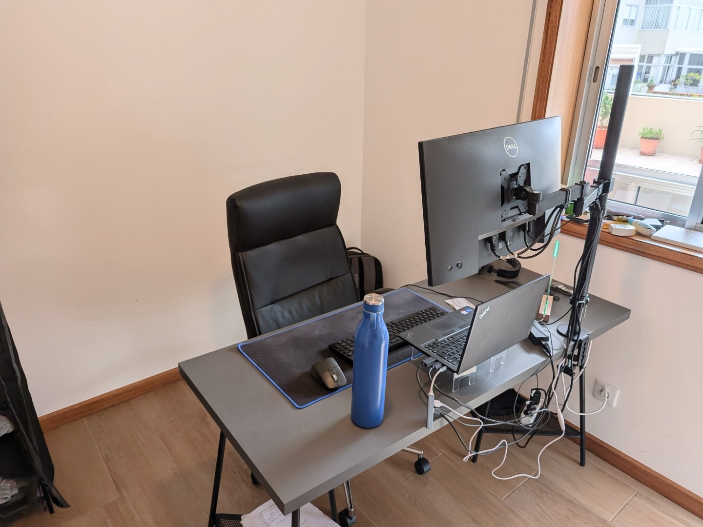Foto de outro ângulo da mesa (por trás), com cadeira e cabos à vista e janela à direita.