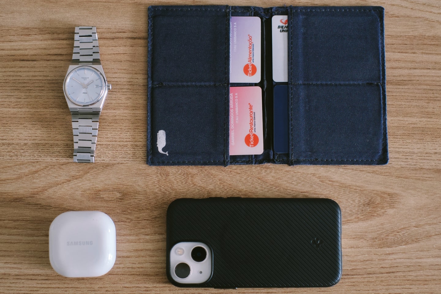 Foto de cima da carteira, relógio, fones de ouvido e celular do Gabriel.