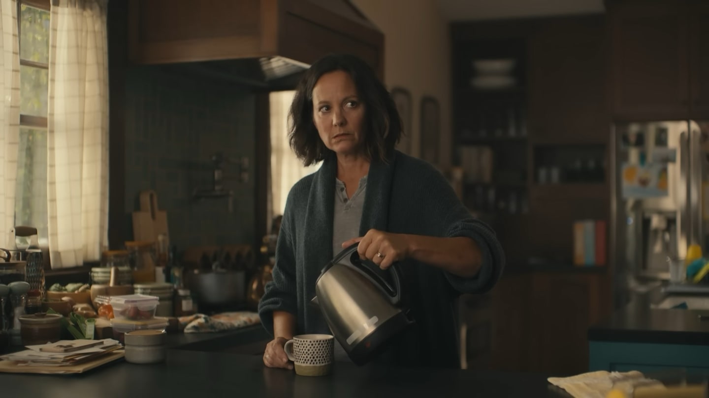 Mulher de meia-idade em uma cozinha, colocando café em uma xícara, olhando para a sua esquerda.