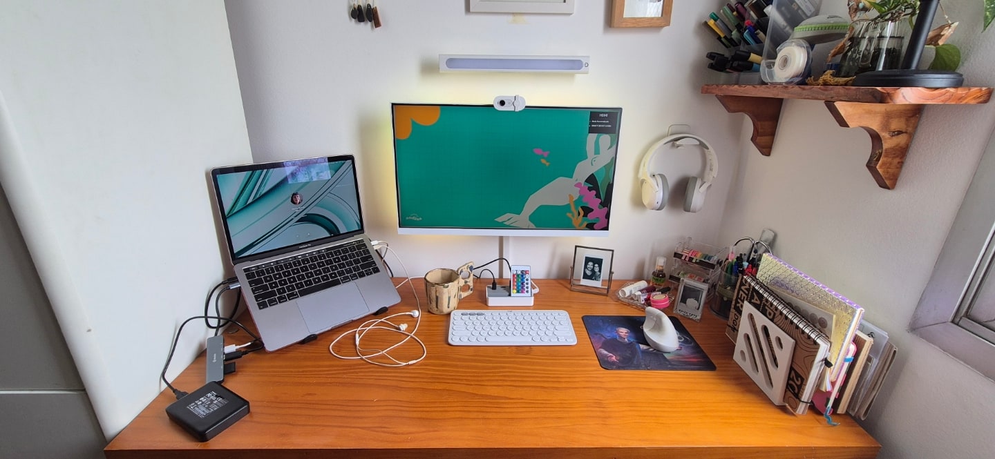 Foto frontal da mesa da Débora, com notebook e monitor no centro, acessórios brancos e uma pequena prateleira suspensa à direita.