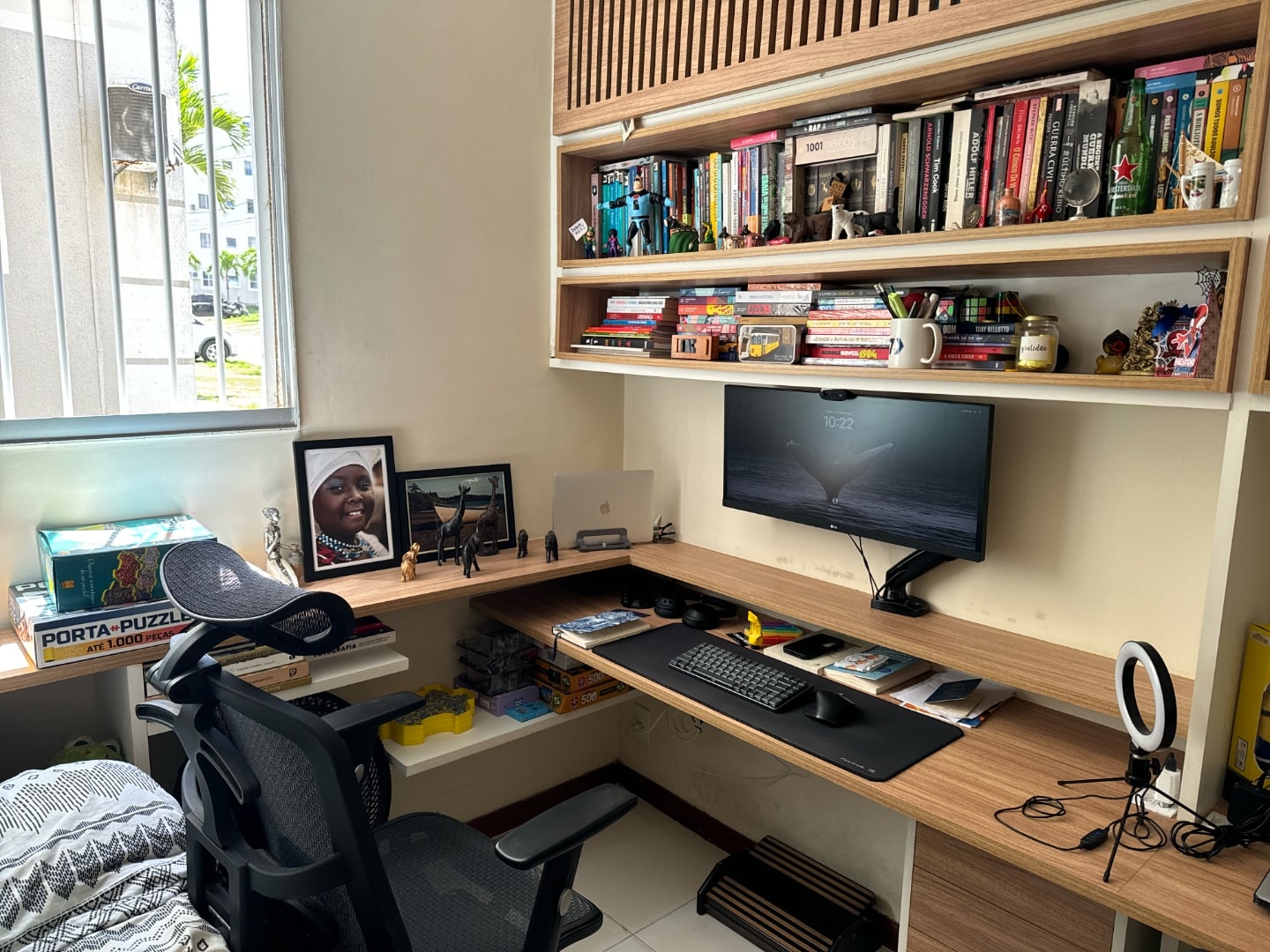 Foto lateral da mesa de trabalho do Tiago, com estante superior, cadeira e objetos de decoração.