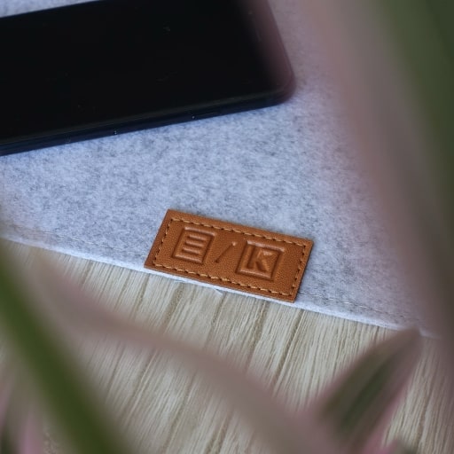 Close da etiqueta em “couro ecológico”, com os logos do Manual do Usuário e da Kumori, no deskmat do Manual, em formato quadrado (1:1).