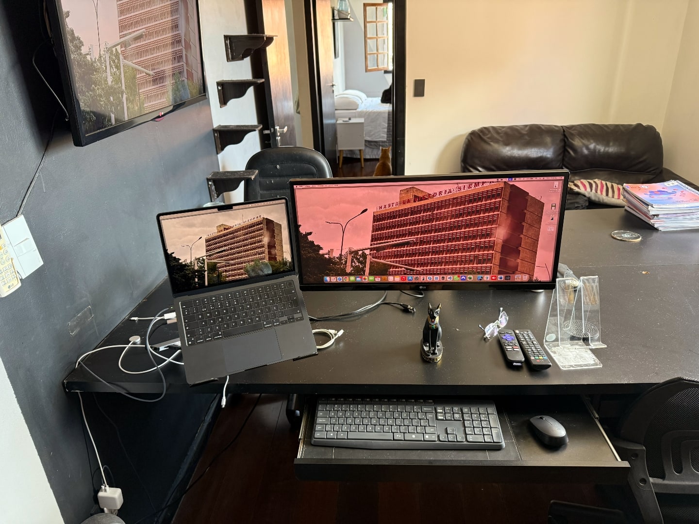 mesa escura, com monitor e notebook, além de acessórios. Ao fundo, sofá de dois lugares e uma TV na parede à esquerda.