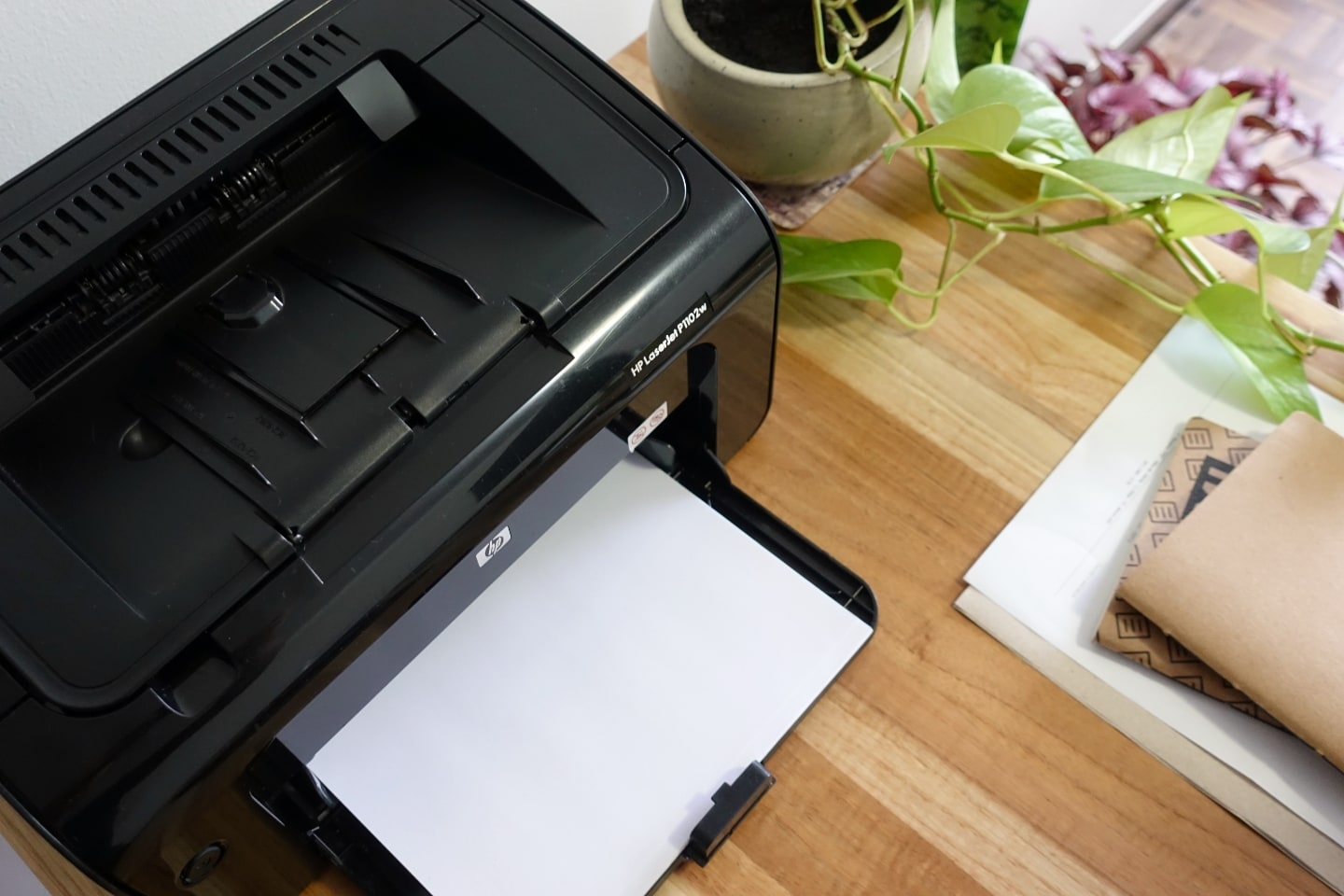 Foto de cima de uma impressora preta, com papel na bandeja, ao lado de um vaso de planta com uma jiboia e alguns papéis e cadernos.